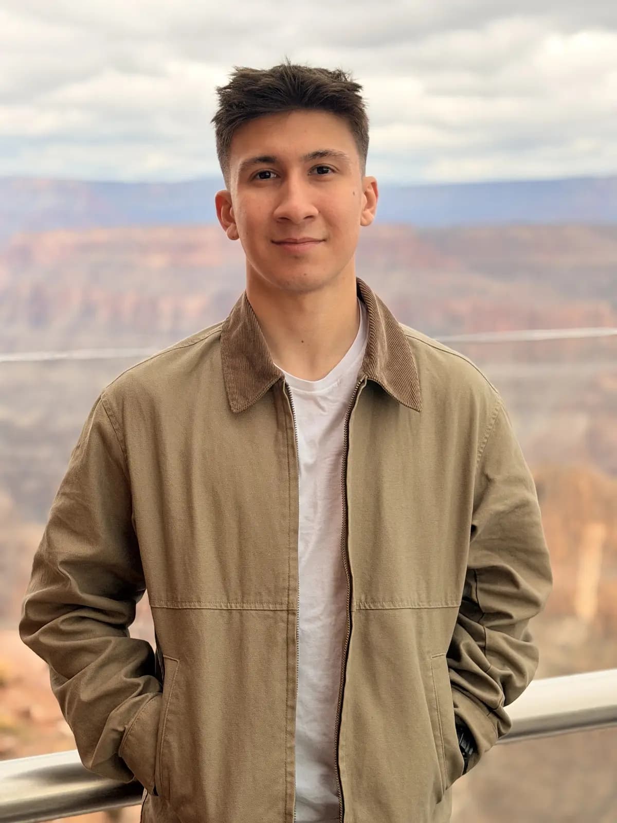 Burak in a tan work jacket and white T-shirt, hands in his pockets, at a railing above the Grand Canyon.