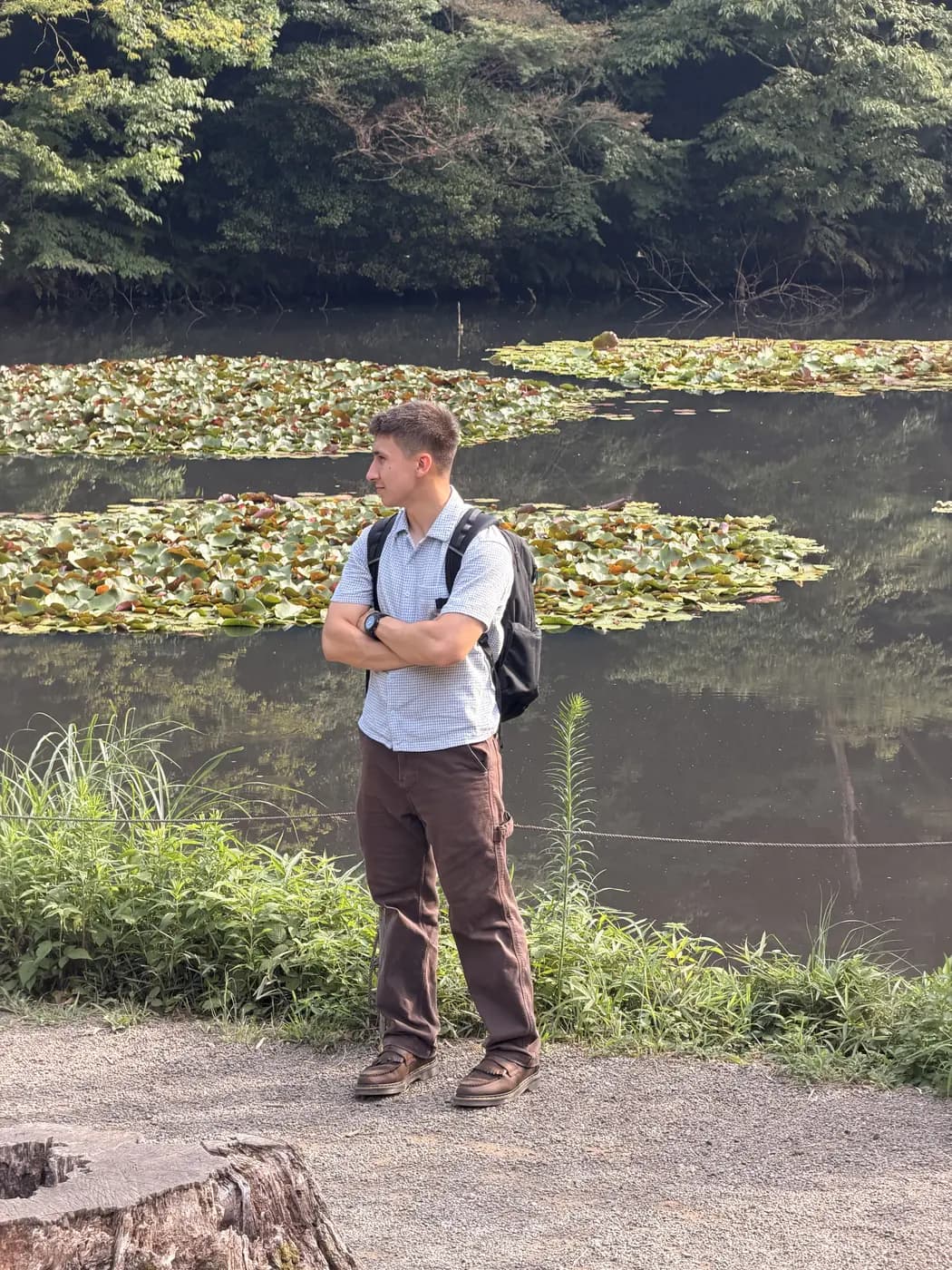 Burak standing on a gravel path beside a pond covered in lily pads, arms folded, wearing a backpack.