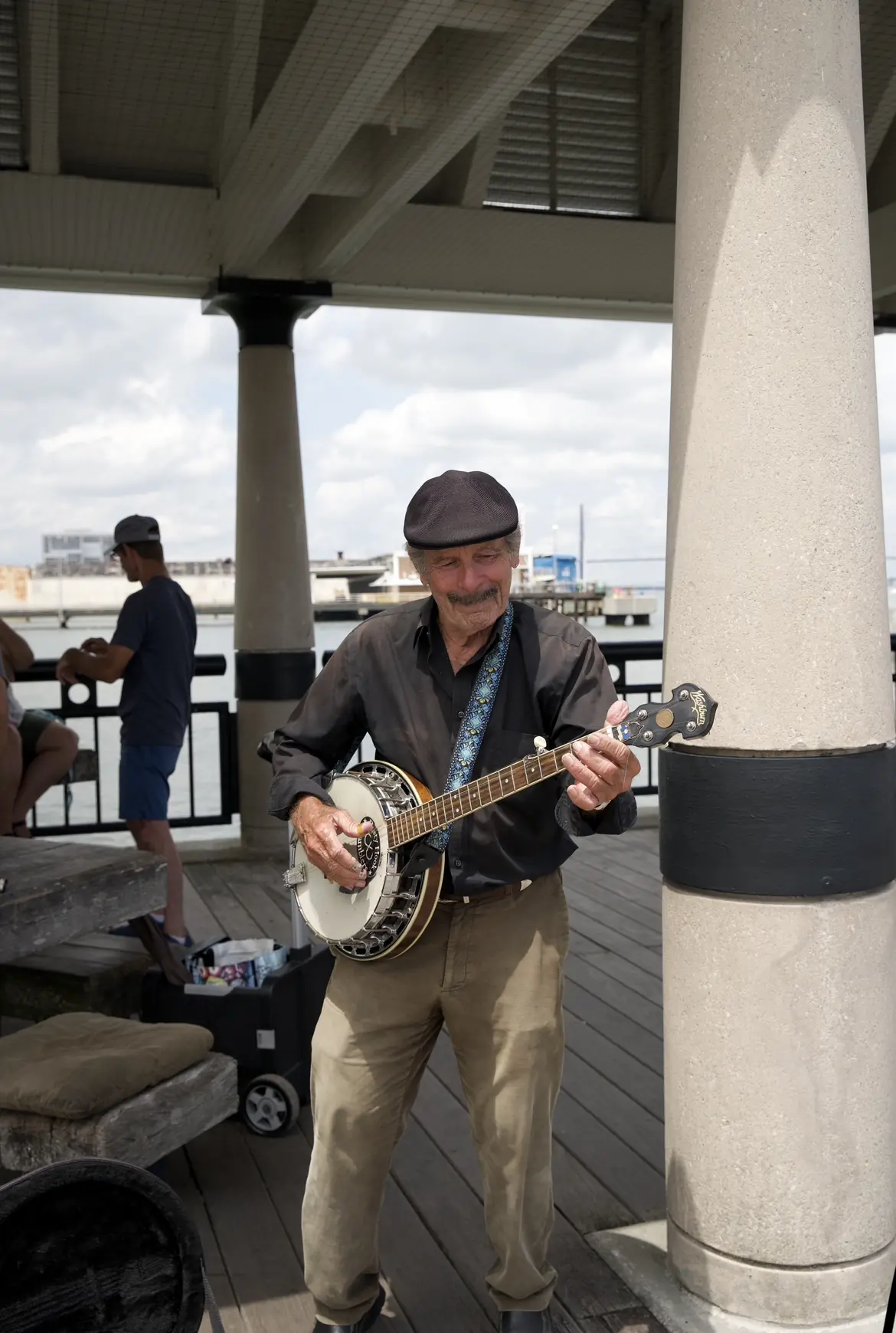 A man in a flat cap playing a banjo under a covered waterfront walkway.