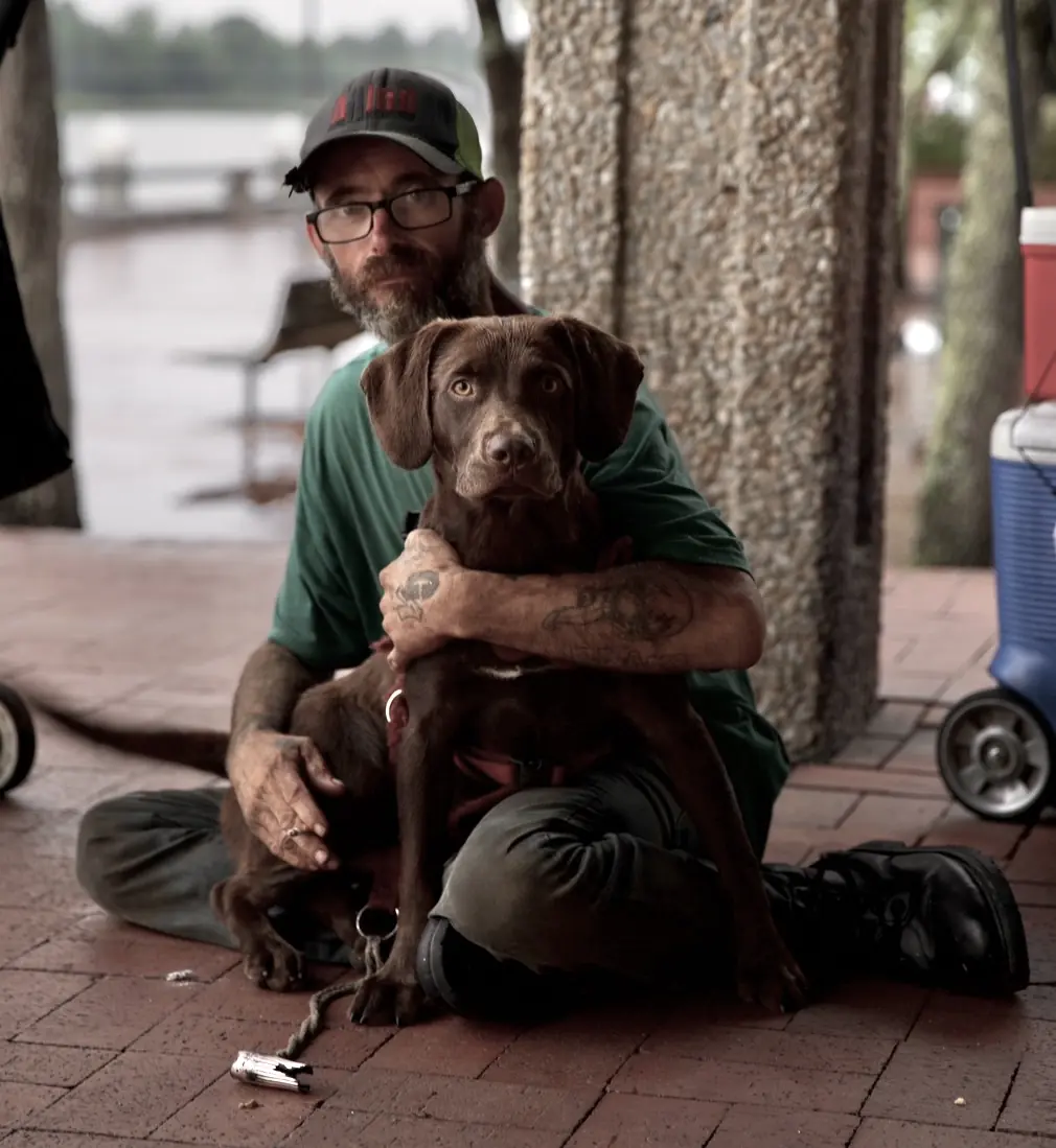 A man sitting on brick paving with his arms around a brown dog.