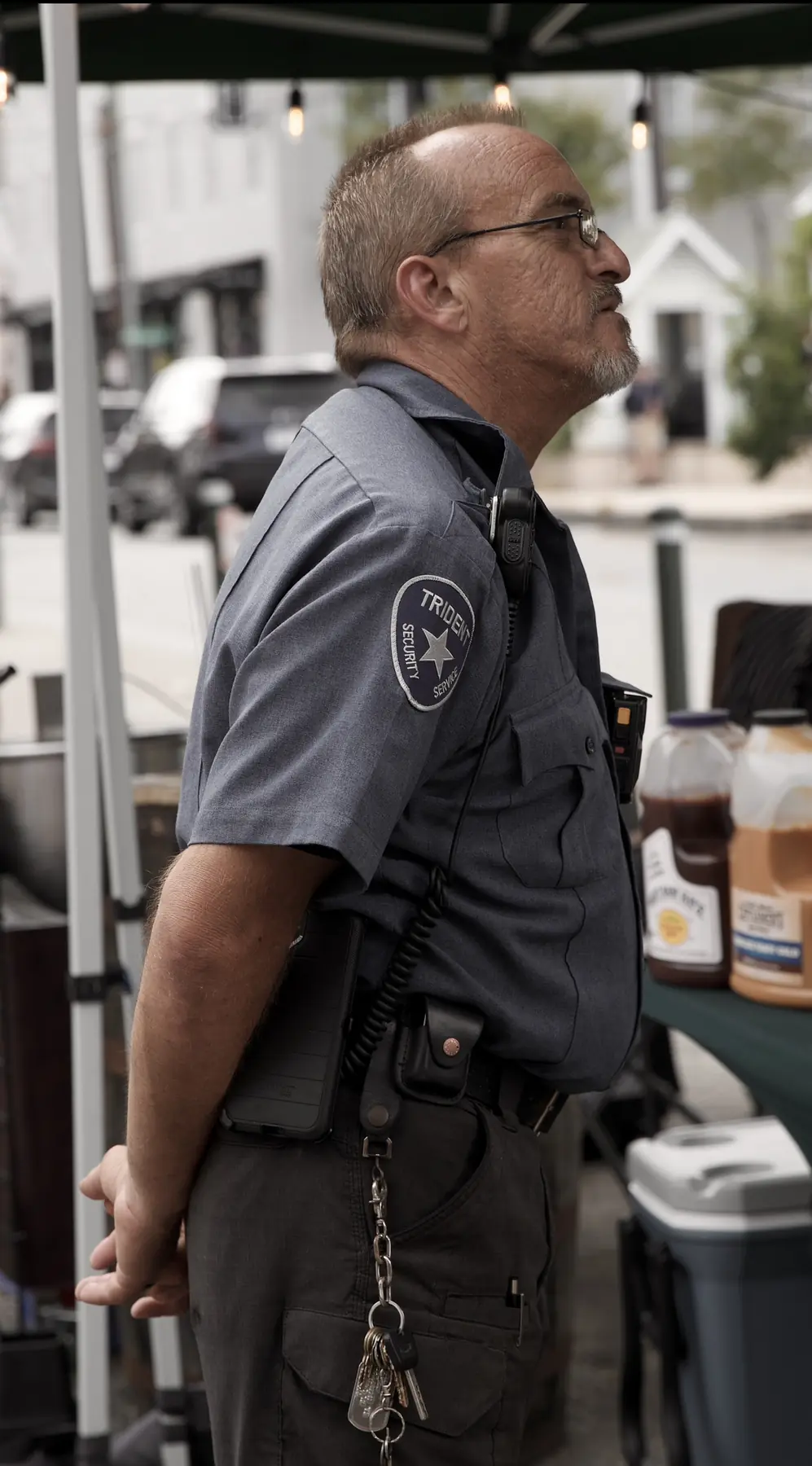 A uniformed security guard in profile beside a market tent.