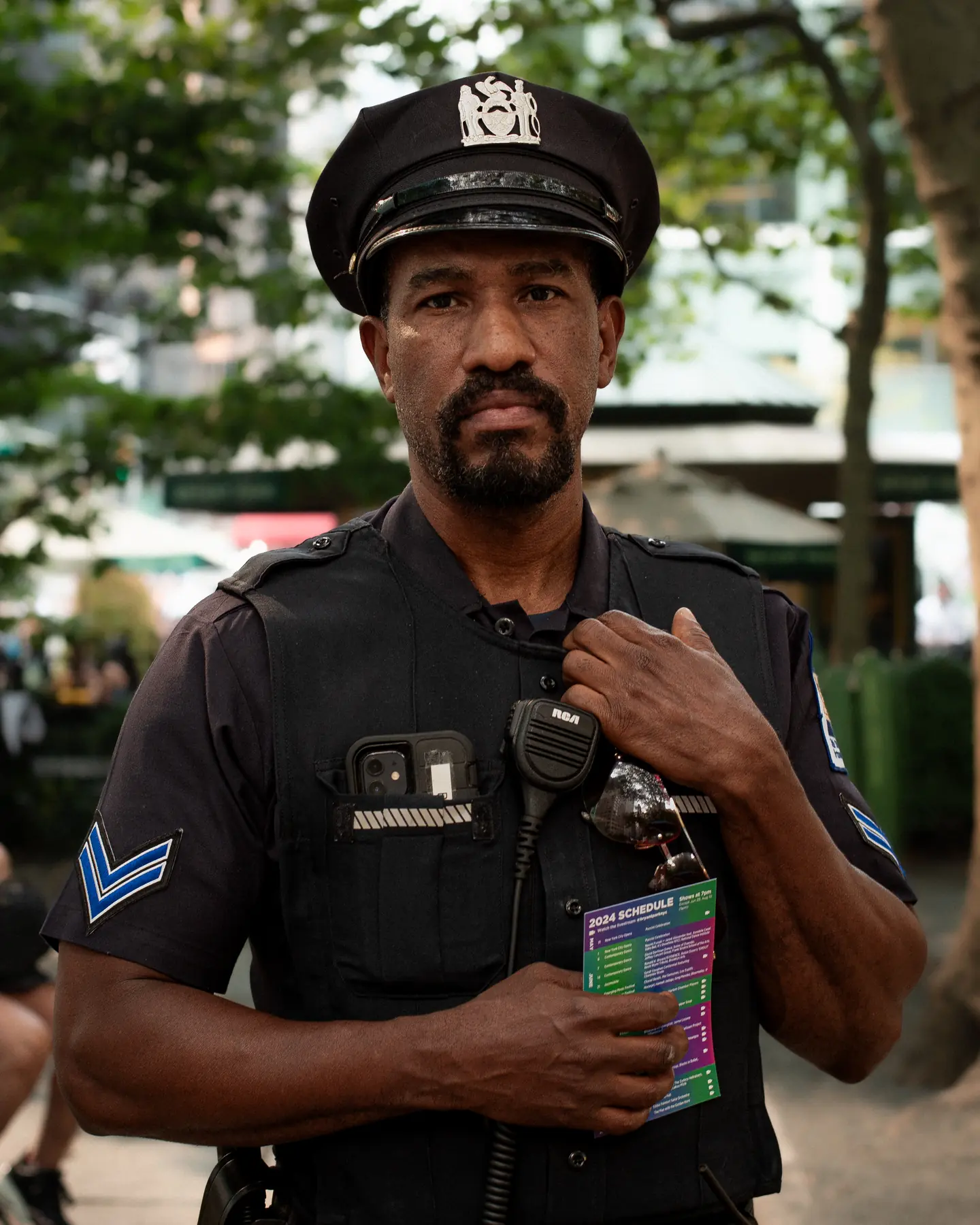 A police officer in uniform and cap, one hand at his chest, under trees.