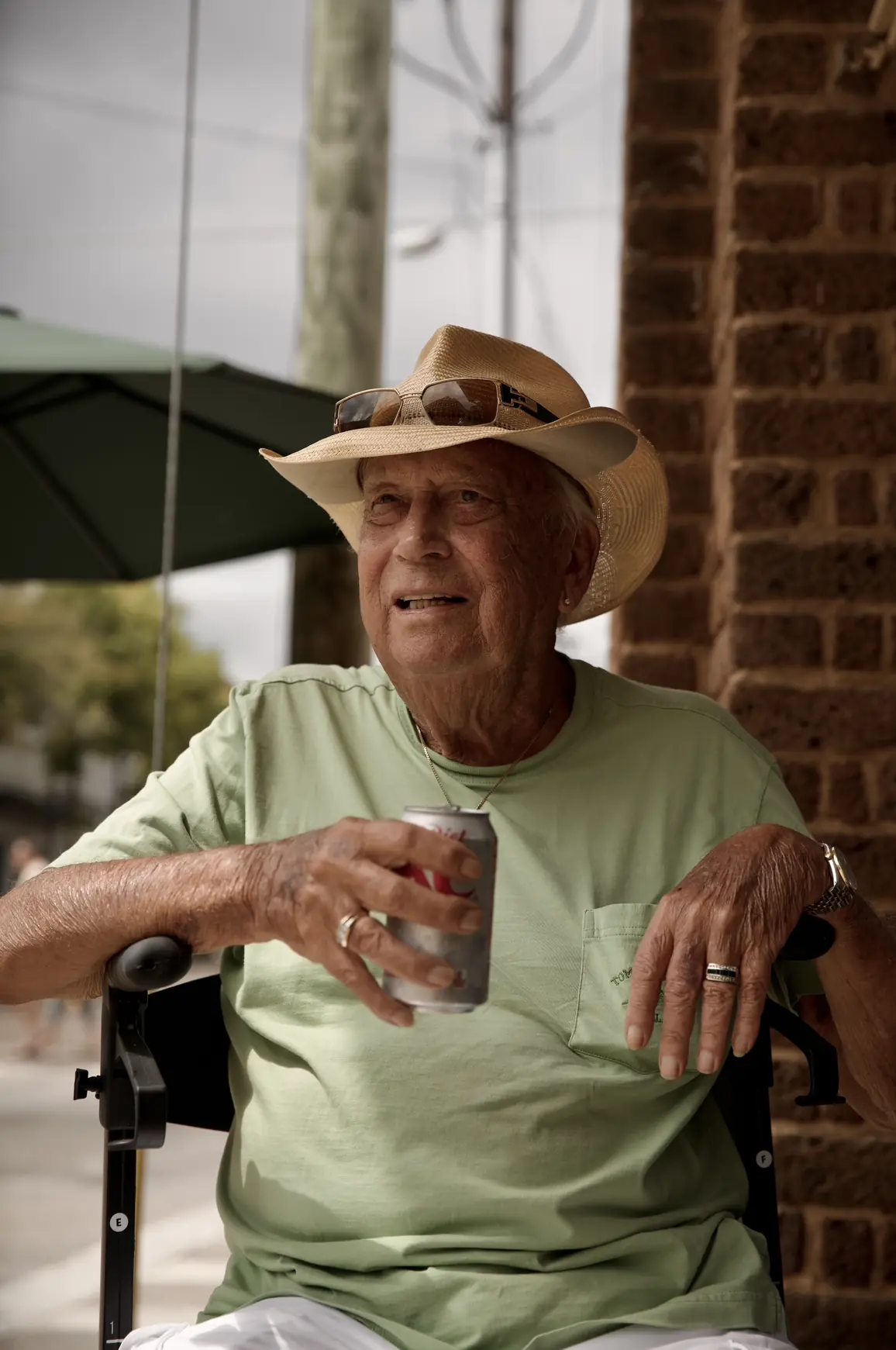 An older man in a cowboy hat seated on a porch, holding a can.