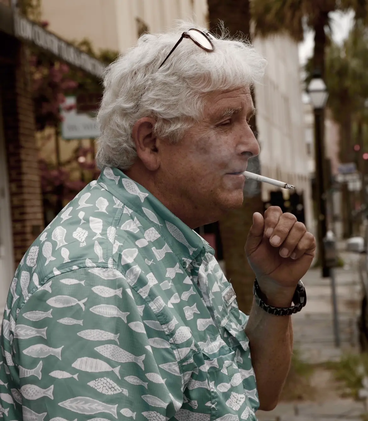 A grey-haired man in a fish-print shirt smoking a cigarette on a street.